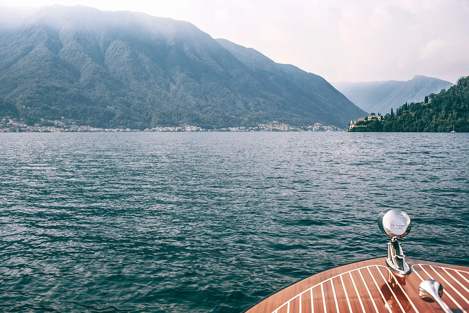 The varnished bow of a wooden boat in the foreground, rippled green water ahead, and Villa del Balbianello on its wooded promontory to the right.