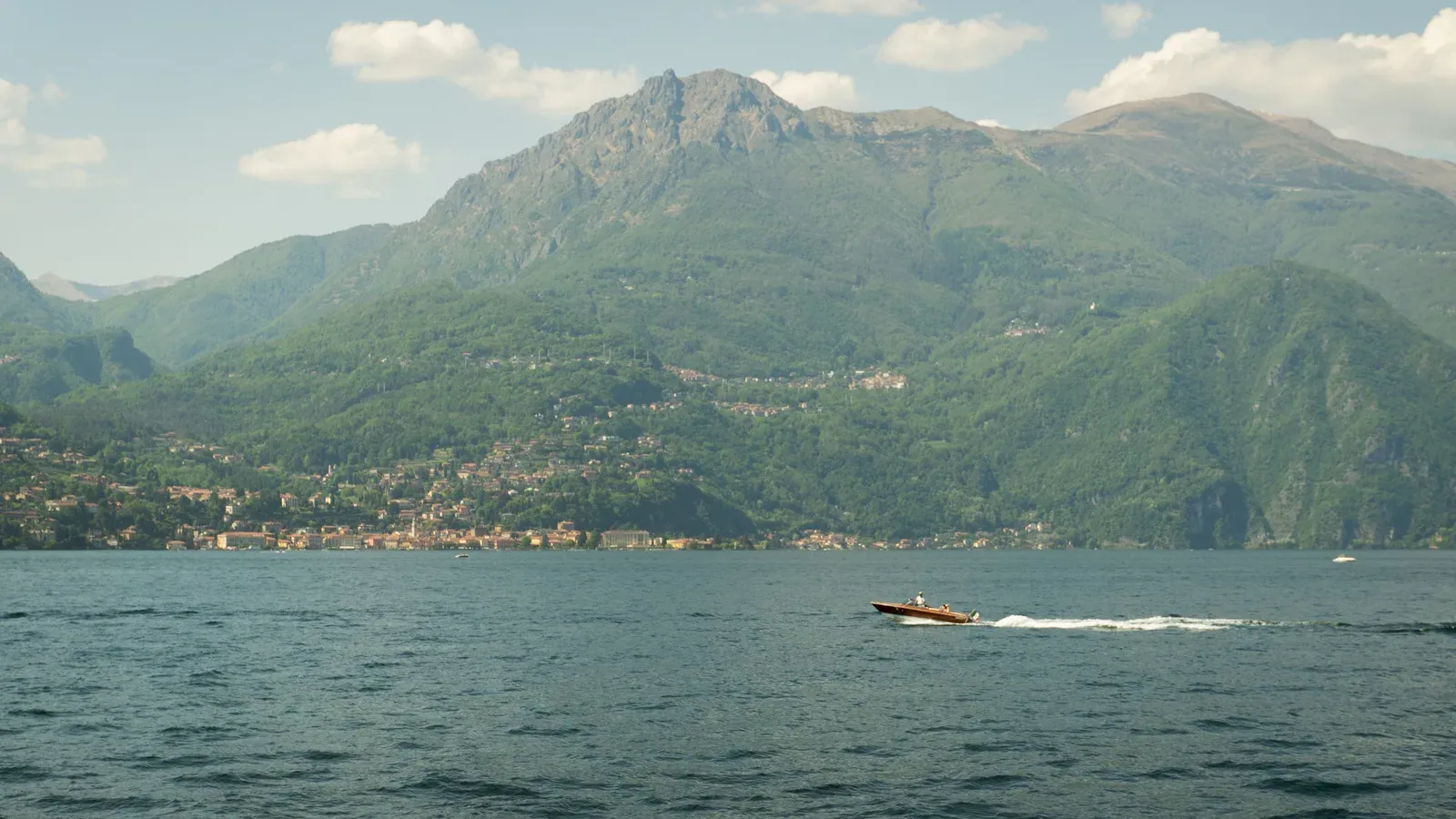 A small varnished wooden motorboat under way on Lake Como with two people aboard, a shoreline town and a rocky green peak behind.