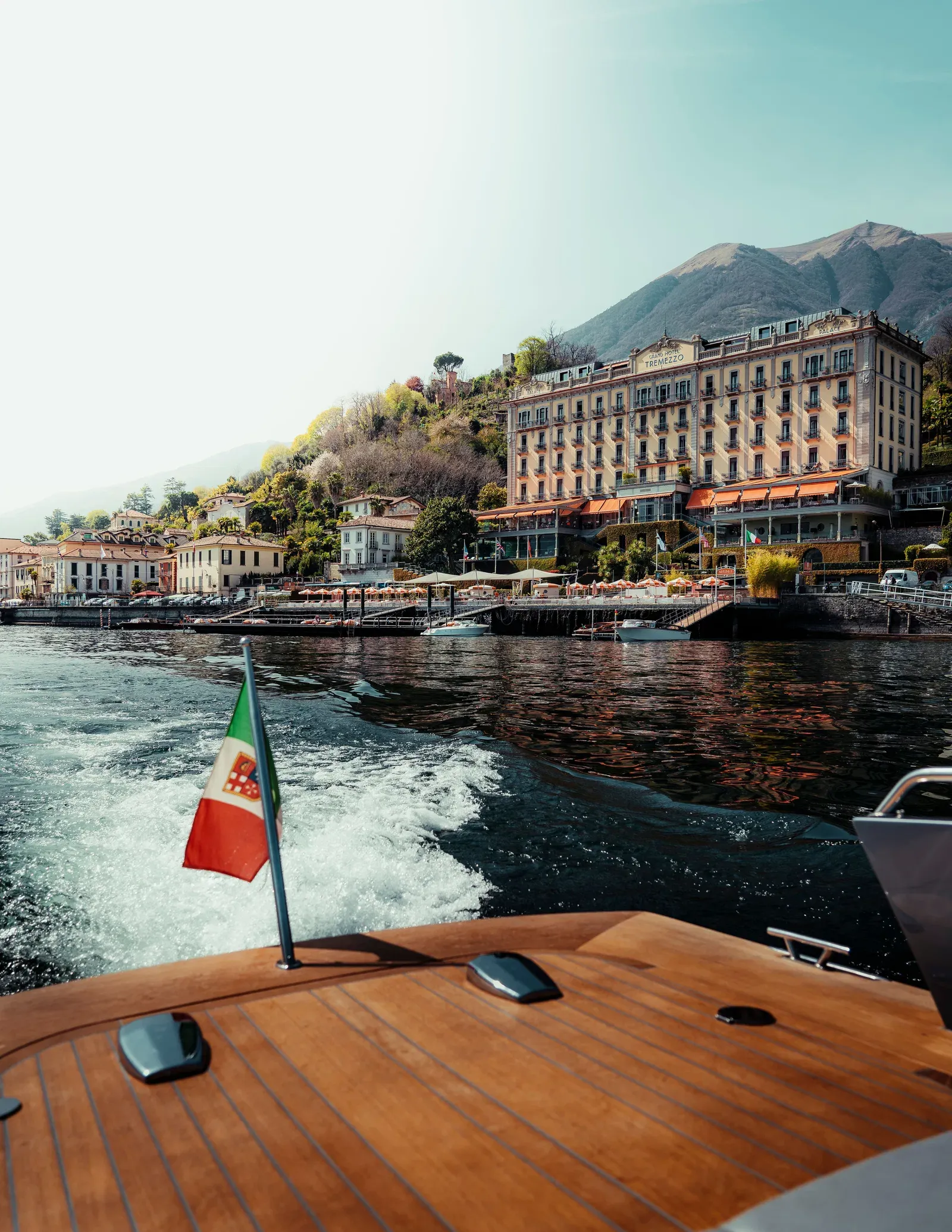 The teak afterdeck of a boat under way, its Italian ensign over the wake, passing a large lakeside hotel with orange awnings in hazy light.
