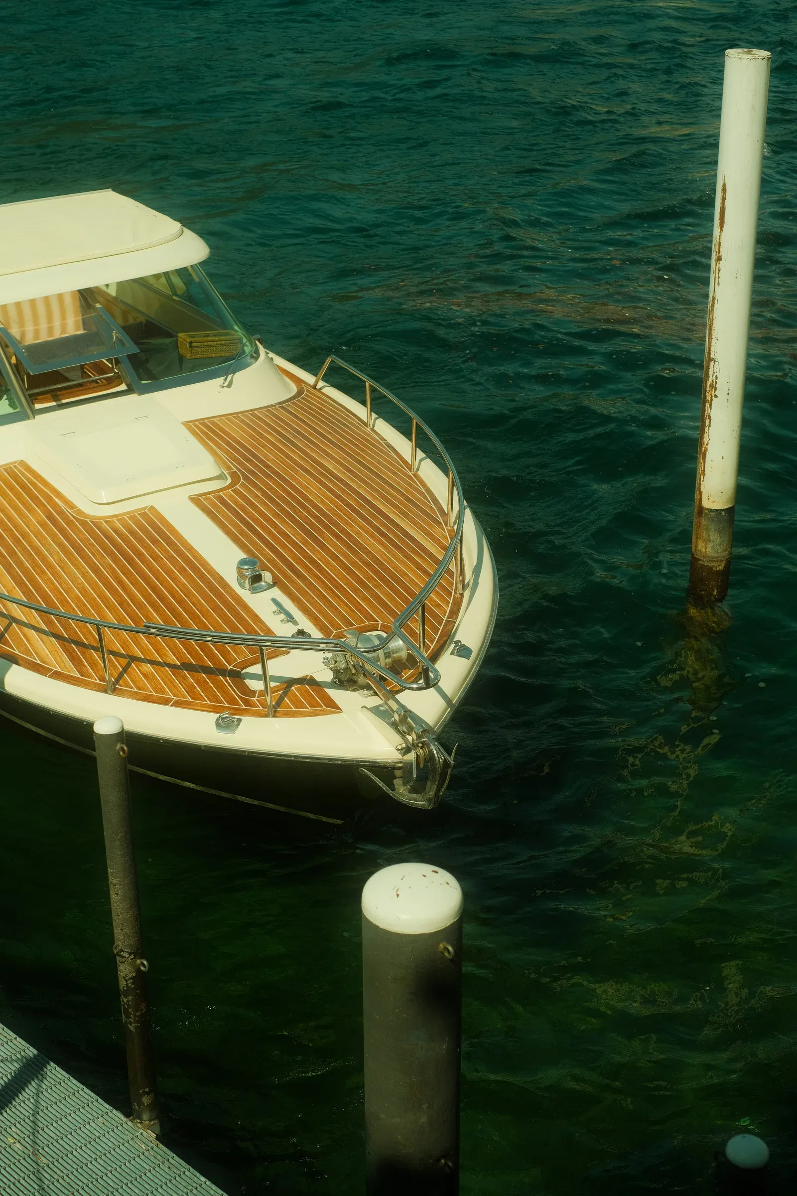 The bow of a white classic motor launch with a varnished teak foredeck, moored beside dock piles in dark green water, seen from above.