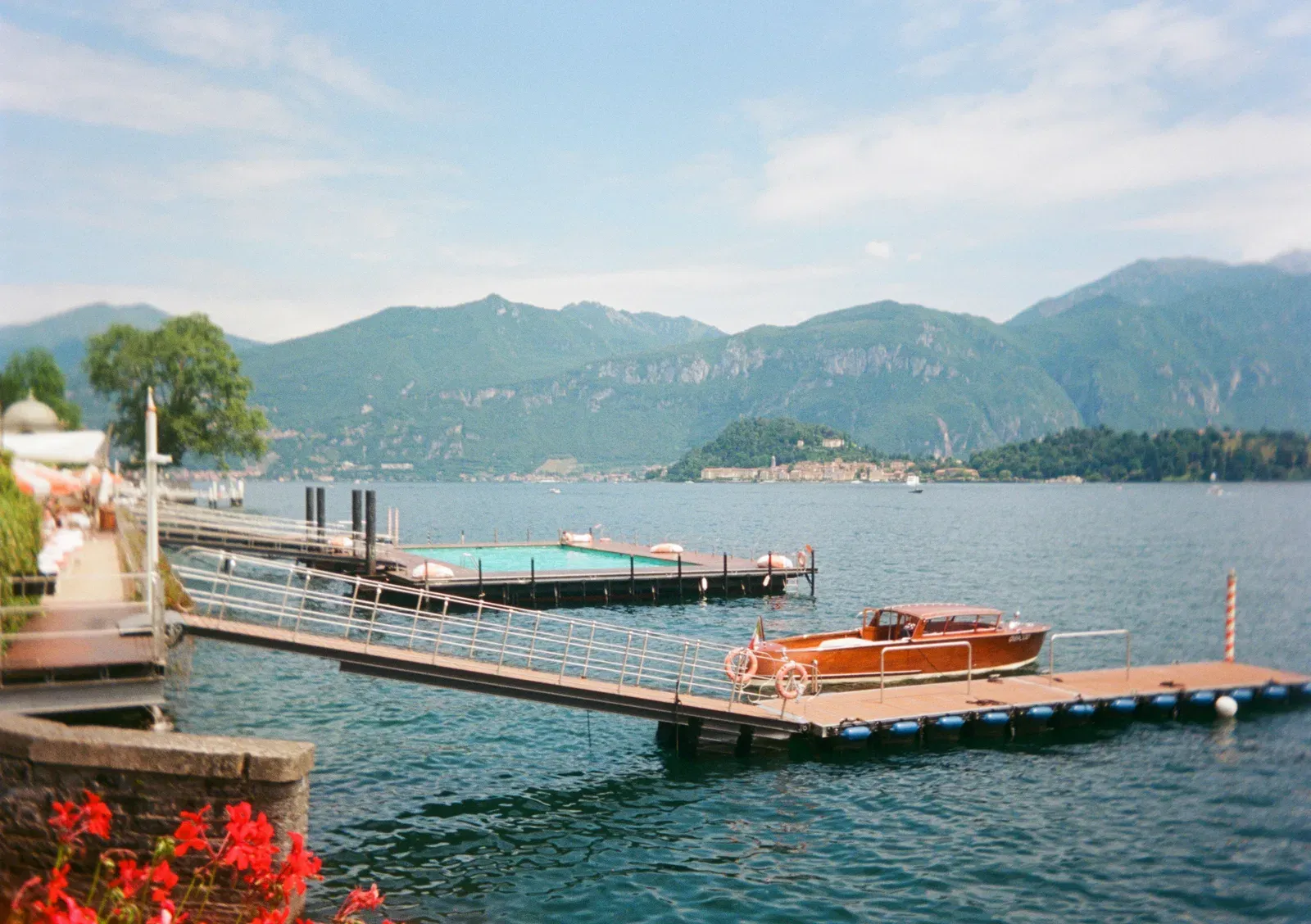 A mahogany taxi boat moored at a floating pontoon beside a lakeside pool deck, hazy summer light, mountains and a far shore town behind.