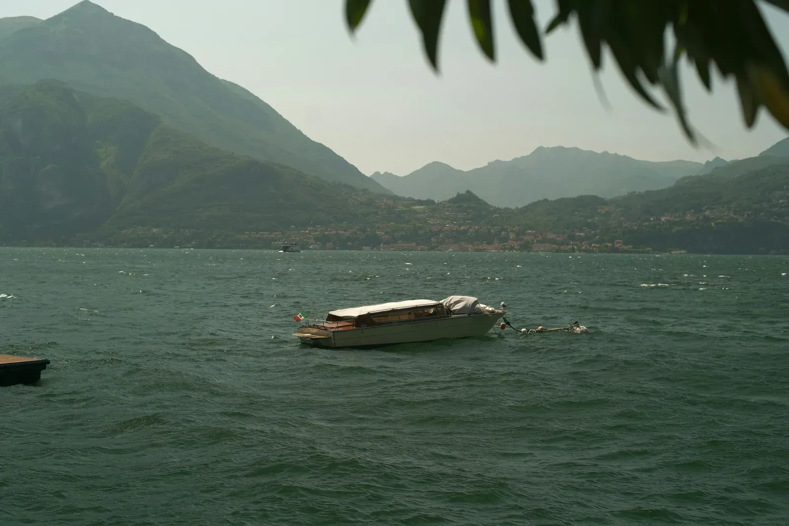A covered white water taxi with an Italian flag, moored to a buoy on choppy dark green water with whitecaps, a ferry and a shoreline town under hazy mountains behind.
