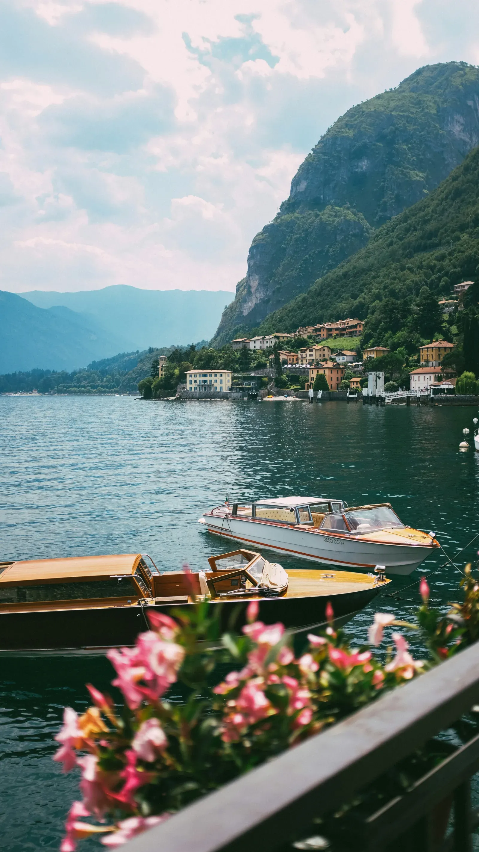 A white water taxi and a varnished wooden runabout moored below a terrace railing with pink oleander, a lakeside village and a wooded cliff on the far shore.