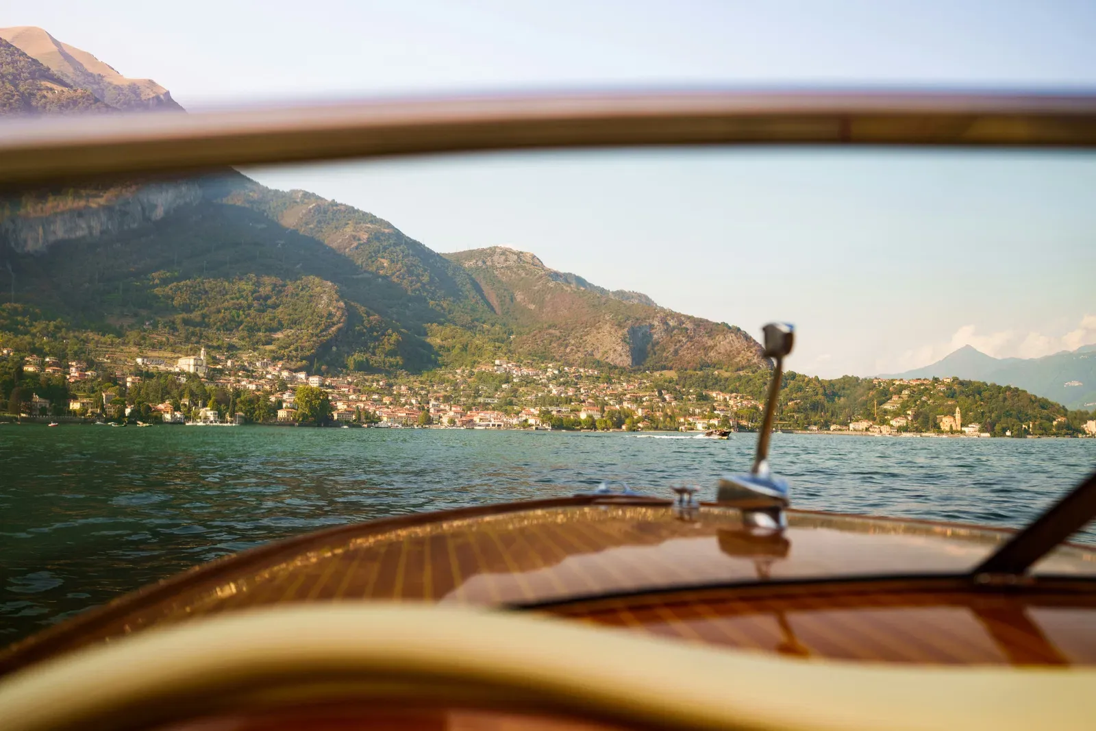The view forward over the varnished mahogany foredeck of a classic motorboat, past the windscreen to a lakeside village and mountains in low, warm light.
