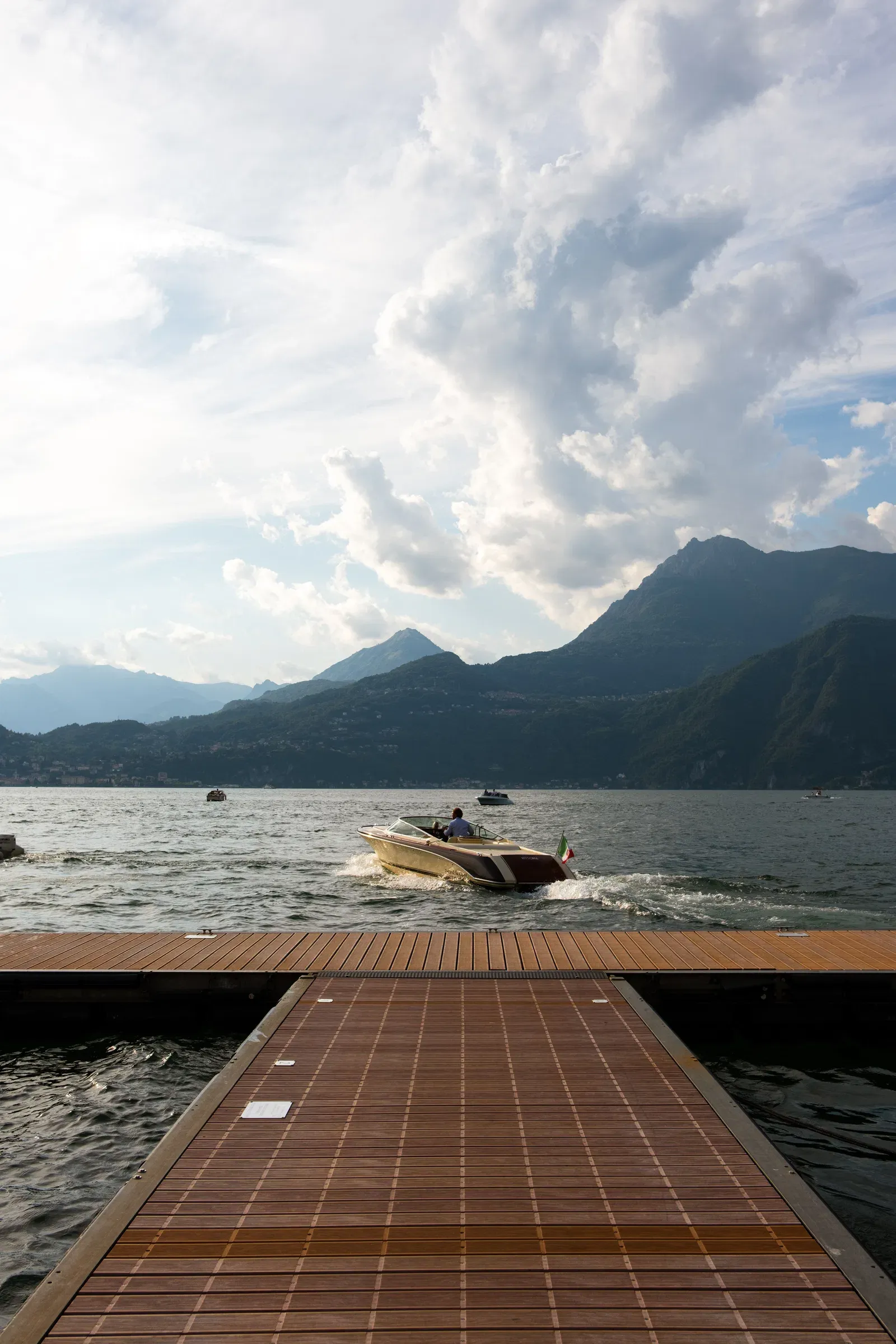 Seen from the end of a wooden pontoon, a classic runabout with two people aboard runs across open water toward mountains under a bank of cloud, in late light.