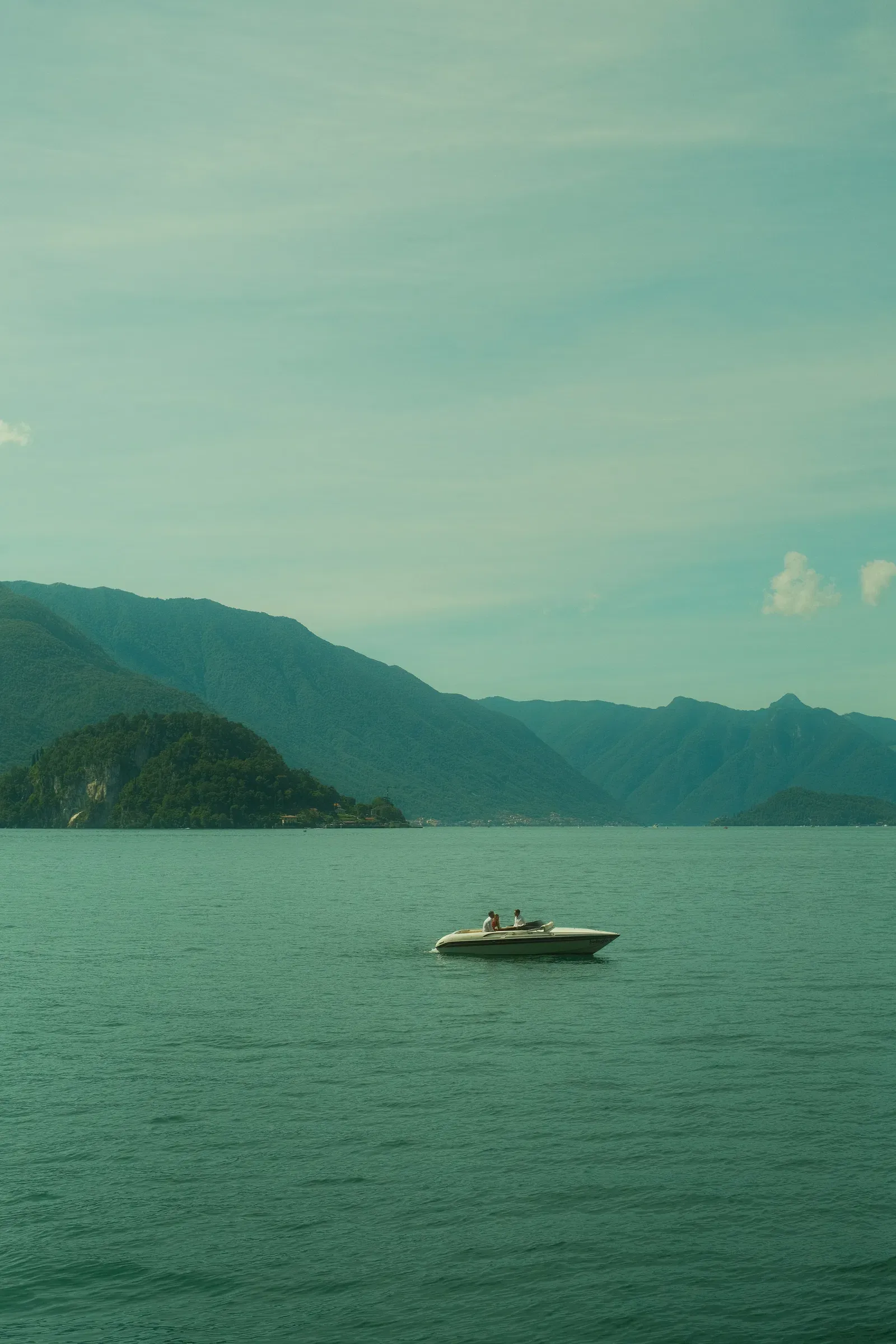 A white sports boat crossing open water on Lake Como with three people aboard, hazy ridges and a wooded headland behind.