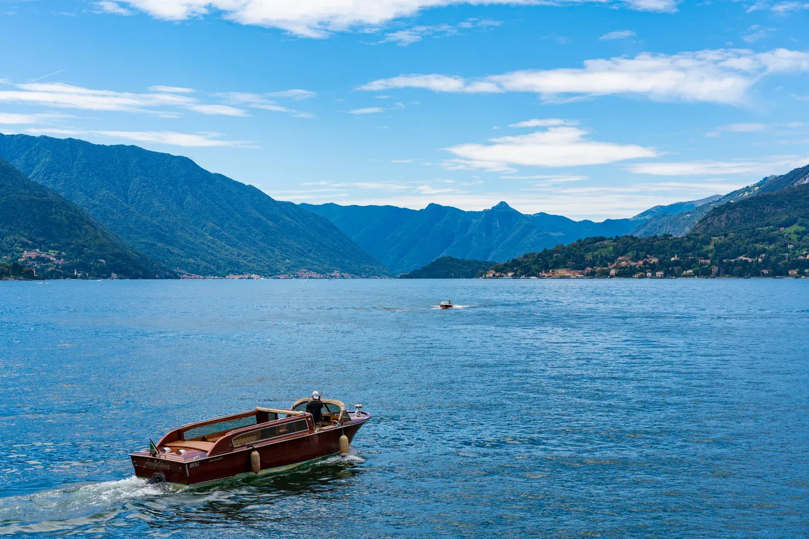 A varnished mahogany taxi boat under way on open water, no passengers aboard, one man standing at the helm, mountains on both shores.