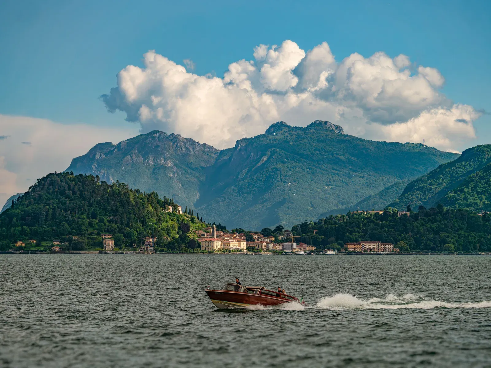 A classic wooden speedboat under way across open water, three people aboard, a lakeside town on a wooded promontory and high mountains behind.